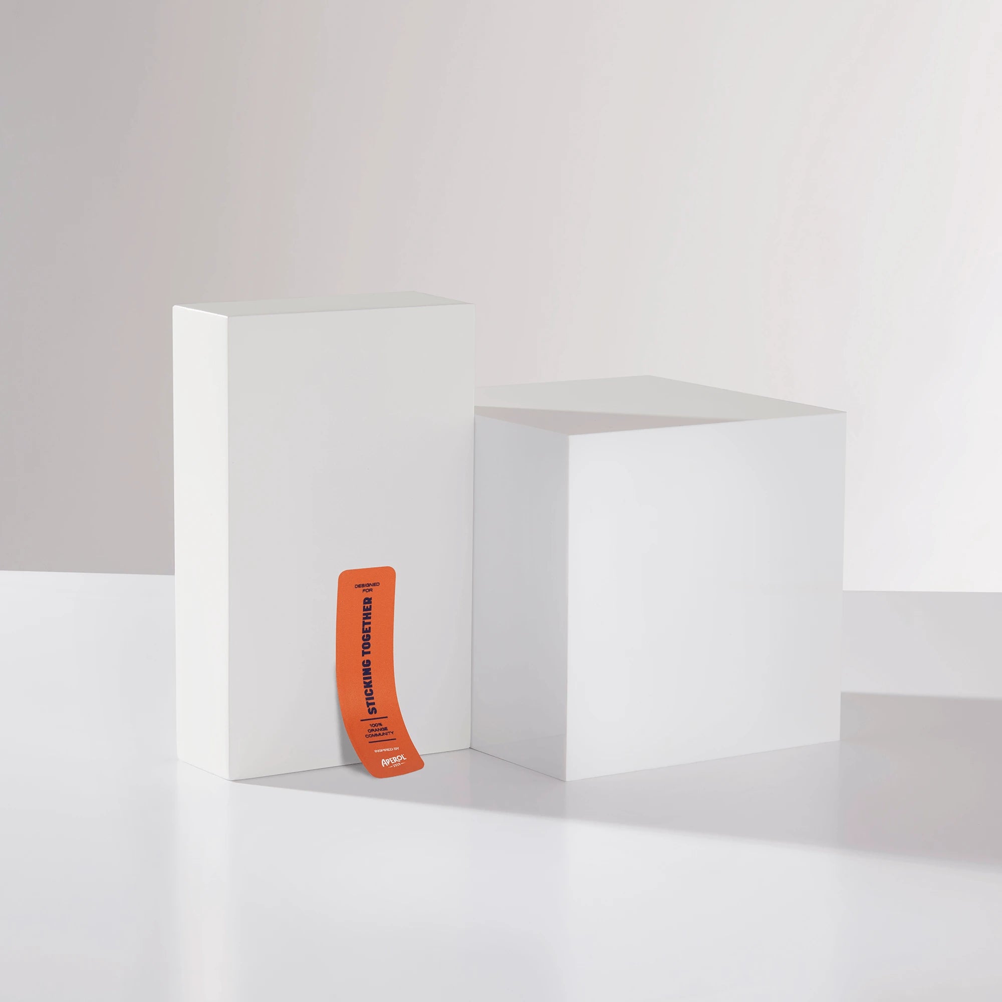 Orange Aperol sticker reading 'STICKING TOGETHER' leaning against minimal white geometric blocks on a clean background.